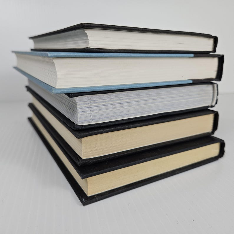 May include: A stack of five hardcover books with black covers and white or cream-colored pages. The top book has a black cover, and the second book has a light blue cover. The books are stacked on a white surface.