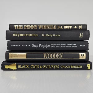 May include: A stack of five hardcover books with black covers and gold lettering. Titles include "THE PENNY WHISTLE", "oxymoronica", "Stay Positive", "WOODEN", and "BLACK CAT'S & EVIL EYES". The books are arranged in a neat pile.