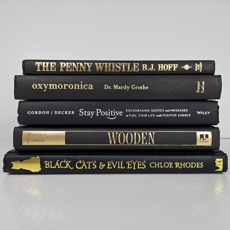 May include: A stack of five hardcover books with black covers and gold lettering. Titles include "THE PENNY WHISTLE", "oxymoronica", "Stay Positive", "WOODEN", and "BLACK CAT'S & EVIL EYES". The books are arranged in a neat pile.