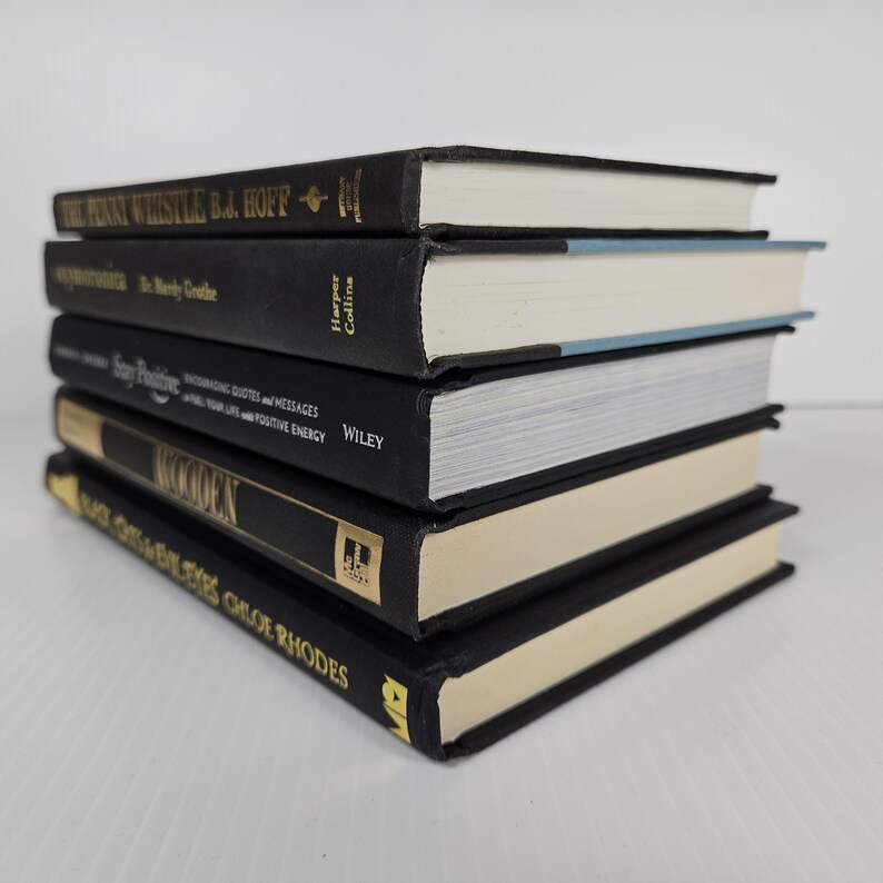 May include: A stack of five hardcover books with black covers and gold lettering. The book titles include "The Funny Whistle," "Stay Positive," and "Wooden." The pages are white, and some have blue or beige edges.