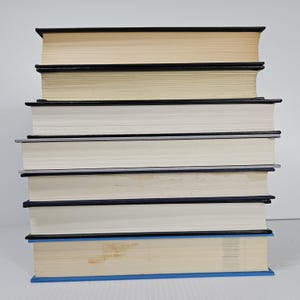 May include: A stack of seven hardcover books with varying colors. The books have black and blue spines, with off-white and cream-colored pages. The books are stacked on top of each other.
