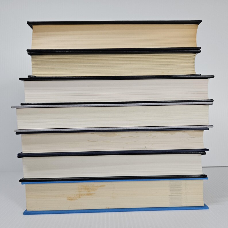 May include: A stack of seven hardcover books with varying colors. The books have black and blue spines, with off-white and cream-colored pages. The books are stacked on top of each other.