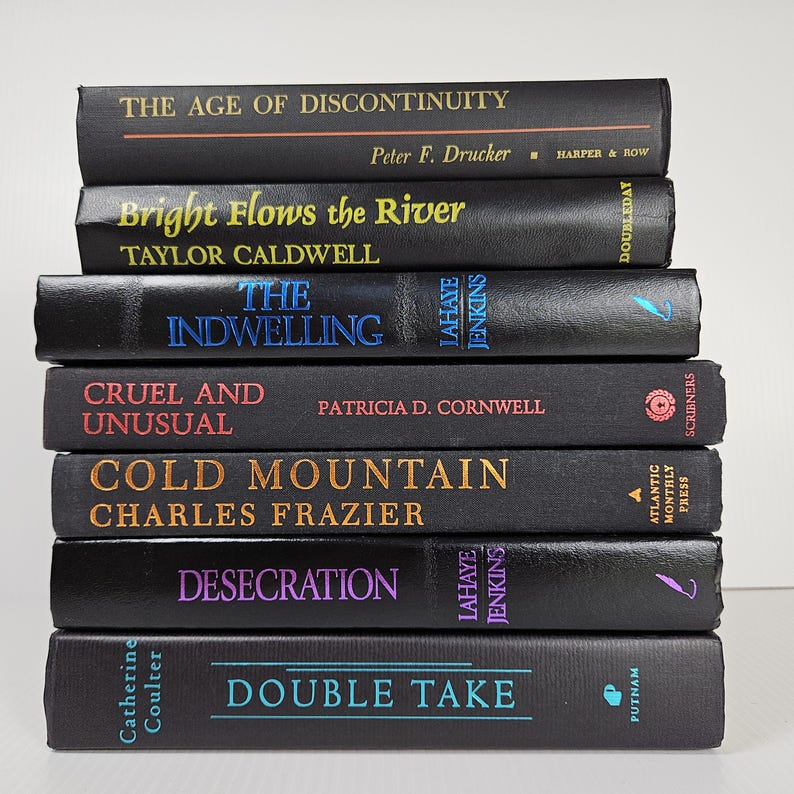 May include: A stack of seven hardcover books with black covers. Titles include "The Age of Discontinuity," "Bright Flows the River," and "The Indwelling." The spines feature author names and publisher imprints in various colors.