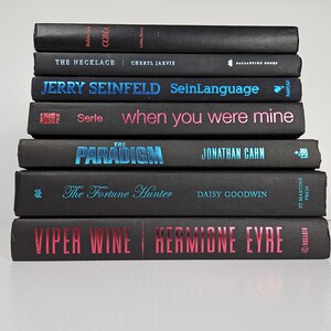 May include: A stack of hardcover books with black covers. Titles include "COMA", "THE NECKLACE", "JERRY SEINFELD SeinLanguage", "when you were mine", "THE PARADIGM", "The Fortune Hunter", and "VIPER WINE". The text is in various colors, including blue and pink.