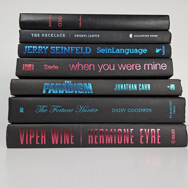 May include: A stack of hardcover books with black covers. Titles include "COMA", "THE NECKLACE", "JERRY SEINFELD SeinLanguage", "when you were mine", "THE PARADIGM", "The Fortune Hunter", and "VIPER WINE". The text is in various colors, including blue and pink.