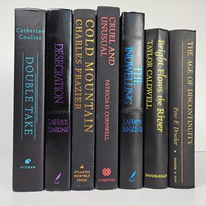 May include: Seven hardcover books with black covers are displayed. Titles include "Double Take," "Desecration," "Cold Mountain," "Cruel and Unusual," "The Indwelling," "Bright Flows the River," and "The Age of Discontinuity." The spines feature author names and publisher imprints.