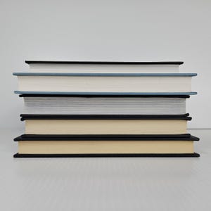 May include: A stack of five hardcover books with varying colors. The books have black spines and edges, with the pages a light cream color. The top book has a black cover, the second a light blue cover, the third a white cover, the fourth a gray cover, and the bottom book a cream cover.