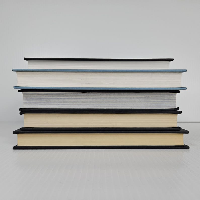 May include: A stack of five hardcover books with varying colors. The books have black spines and edges, with the pages a light cream color. The top book has a black cover, the second a light blue cover, the third a white cover, the fourth a gray cover, and the bottom book a cream cover.