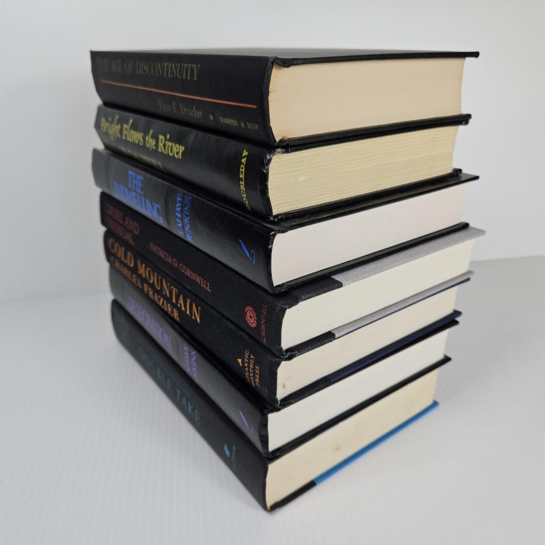 May include: A stack of seven hardcover books with black covers and white pages. Titles include "The Age of Discontinuity", "Bright Flows the River", and "Cold Mountain". The books are arranged in a neat pile, with the spines facing the viewer.
