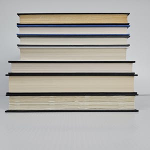 May include: A stack of hardcover books with black spines and cream-colored pages. The top book has a gold-colored top edge, followed by a blue book, and then several books with white pages. The books are neatly arranged, creating a visually appealing composition.