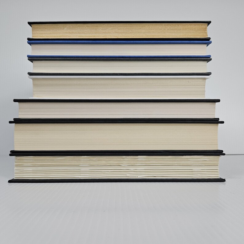 May include: A stack of hardcover books with black spines and cream-colored pages. The top book has a gold-colored top edge, followed by a blue book, and then several books with white pages. The books are neatly arranged, creating a visually appealing composition.