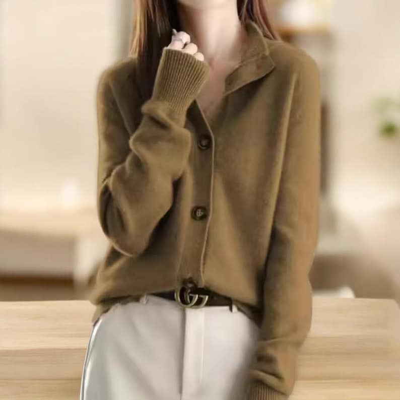 May include: A button-down cardigan in a muted olive green color. The sweater has a ribbed cuff and a collar. The buttons are a darker shade of brown. The person is wearing a black belt with a silver buckle and white pants.