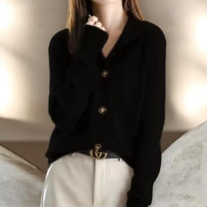 May include: A black cardigan with a collared neckline and three round, brown buttons. The long-sleeved sweater is worn with a black belt with a gold buckle and white pants. The cardigan is a versatile piece of clothing.