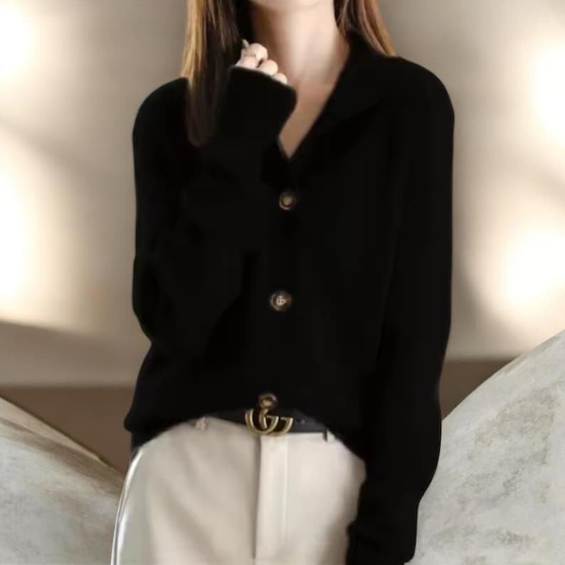 May include: A black cardigan with a collared neckline and three round, brown buttons. The long-sleeved sweater is worn with a black belt with a gold buckle and white pants. The cardigan is a versatile piece of clothing.