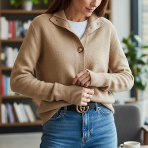 May include: A tan cardigan with a button closure, worn over a white t-shirt. The cardigan has a collared neckline and ribbed cuffs. The person is wearing blue jeans and a black belt with a gold buckle. A bookshelf is in the background.