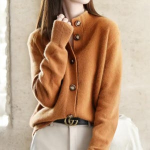 May include: A burnt orange cardigan with a high collar and three brown buttons. The long-sleeved sweater has ribbed cuffs and a matching ribbed collar. The cardigan is paired with a black belt and light-colored pants.