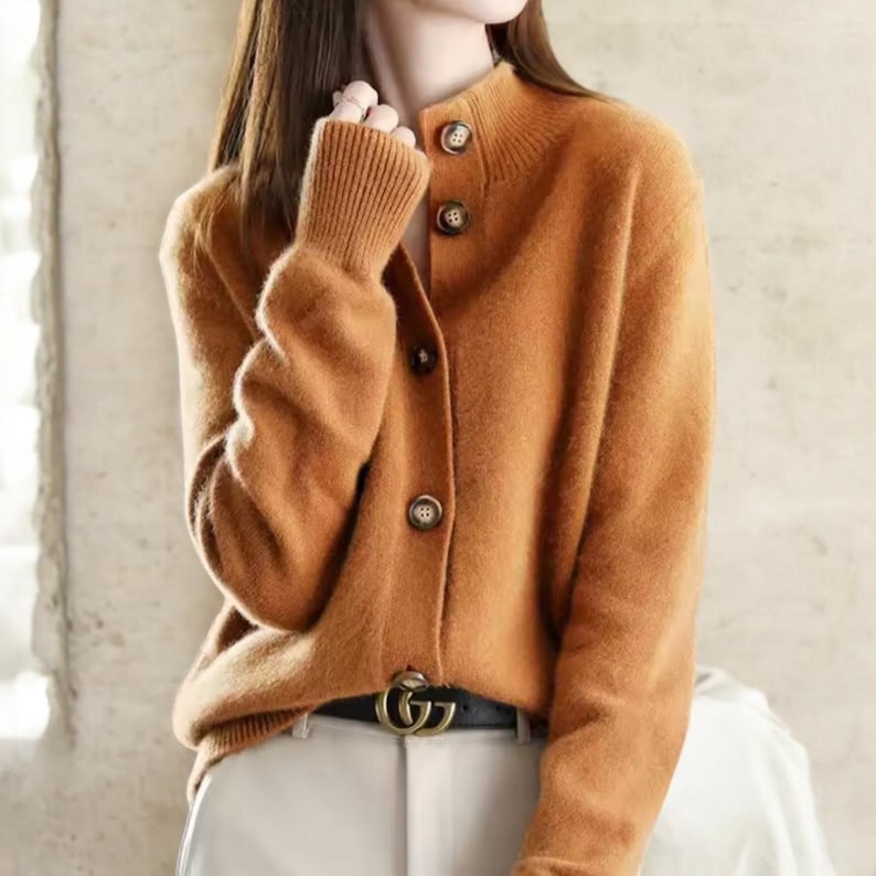 May include: A burnt orange cardigan with a high collar and three brown buttons. The long-sleeved sweater has ribbed cuffs and a matching ribbed collar. The cardigan is paired with a black belt and light-colored pants.