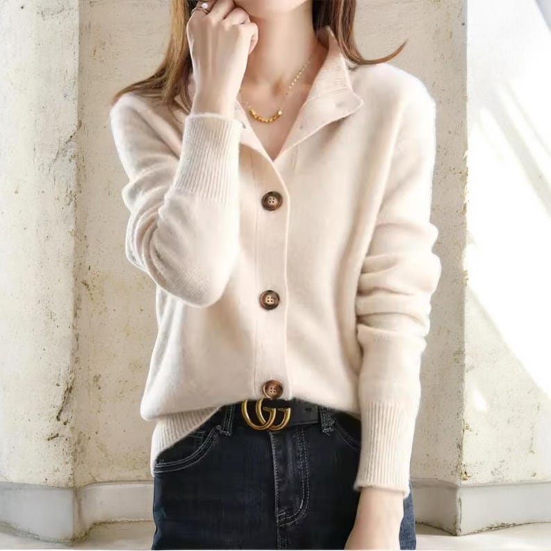 May include: Cream-colored cardigan sweater with a collared neckline and three brown buttons. The long-sleeved sweater has ribbed cuffs and is paired with dark blue jeans and a gold-colored belt.