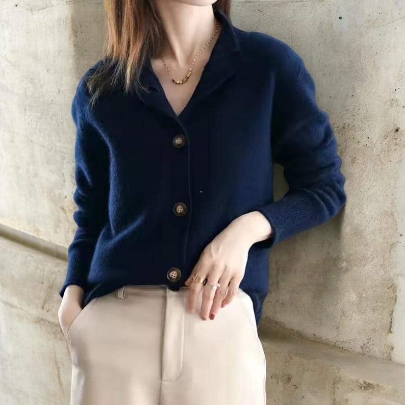 May include: A navy blue cardigan with a collared neckline and three brown buttons. The long-sleeved sweater is paired with beige pants. A gold necklace is visible.