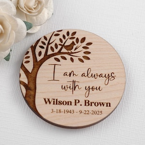 May include: A round wooden memorial keepsake with a tree and bird design. The text "I am always with you" is engraved, along with a name and dates. The wood has a light color with a darker brown engraving.