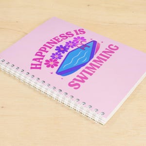 Könnte beinhalten: Ein spiralgebundenes Notizbuch mit hellrosa Einband. Auf dem Cover steht in Retro-Schrift "Happiness is Swimming", zusammen mit einer Grafik eines Schwimmbeckens und Blumen. Das Notizbuch liegt auf einer hellen Holzoberfläche. Ca. 20 cm lang.