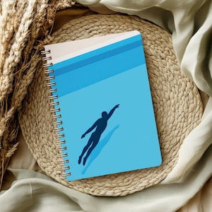Könnte beinhalten: Spiralgebundenes Notizbuch mit blauem Einband, der eine Schwimmer-Silhouette zeigt. Das Design umfasst horizontale blaue Streifen und einen Schatteneffekt. Das Notizbuch liegt auf einer gewebten Matte. Ca. 20 cm lang.