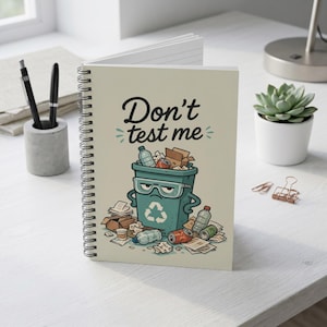 May include: Spiral-bound notebook with a cover featuring a cartoon trash can overflowing with recyclables. The text "Don't test me" is above the trash can. The notebook is open, revealing blank pages. Pens and a succulent plant are in the background.