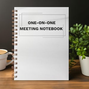 May include: A white spiral-bound notebook with the text "ONE-ON-ONE MEETING NOTEBOOK" printed on the cover. A white coffee cup with a gold rim and a small potted plant are on the wooden surface.