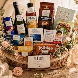 Op de afbeelding: Een gevlochten rieten geschenkmand gevuld met diverse etenswaren en dranken, waaronder een fles mousserende wijn, Red Bull, koffie, chocolade en snacks. Elk item is voorzien van een klein wit label met Duitse tekst. De mand is versierd met kleine roze bloemen en een groot bord aan de voorzijde met de tekst ELTERN SURVIVAL KIT.