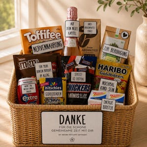Op de afbeelding: Een gevlochten mand gevuld met diverse snacks en een fles mousserende wijn. De items omvatten Toffifee, M&Ms, Snickers Minis, Haribo Goudberen, Extra kauwgom, Hitschies, Dextro Energy, Tchibo koffie en kruidenthee, elk met kleine witte labels.