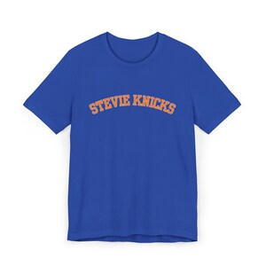 Peut inclure: Un t-shirt bleu roi à manches courtes avec le texte STEVIE KNICKS imprimé en arc de cercle dans une police de style universitaire sur la poitrine. Les lettres sont orange avec un contour blanc, créant un contraste net sur le tissu bleu.