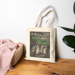 May include: Cream-colored tote bag with a rectangular graphic featuring the text "I'M WAITING FOR IT THAT GREEN LIGHT I WANT IT" in green and white. The graphic also includes a vintage image of people dancing. The bag has two handles.