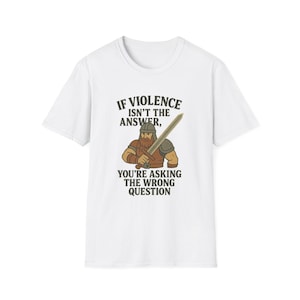 Puede incluir: Camiseta blanca con una ilustración de un vikingo sosteniendo una espada. El texto dice: "Si la violencia no es la respuesta, estás haciendo la pregunta equivocada". El diseño utiliza colores marrón, beige y negro.