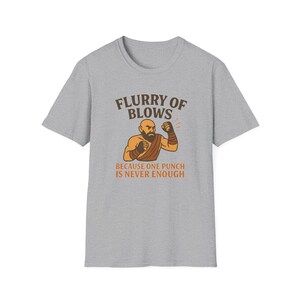 May include: A gray t-shirt featuring a cartoon illustration of a muscular man in a fighting stance. The text above the illustration reads "Flurry of Blows," and below it, "Because One Punch Is Never Enough."