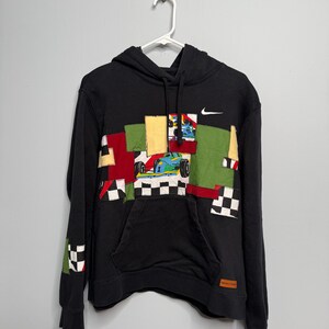 May include: Black hoodie with a patchwork design featuring race cars, checkered flags, and geometric shapes in red, green, yellow, and white. The Nike logo is visible on the upper right chest.