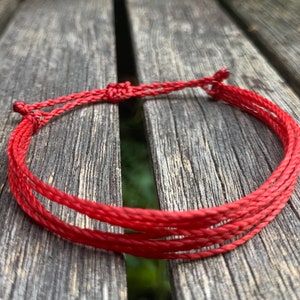 May include: A red braided bracelet made of multiple strands of cord. The bracelet is tied in a knot at the end.