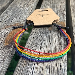 May include: Rainbow colored string bracelet with black cord ends. The bracelet is on a wooden surface.
