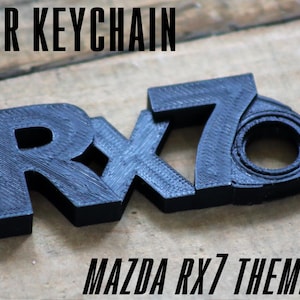 May include: Black Mazda RX7 themed car keychain. The keychain is made of a textured material and features the RX7 logo. The words "CAR KEYCHAIN" and "MAZDA RX7 THEMED" are printed on the image.