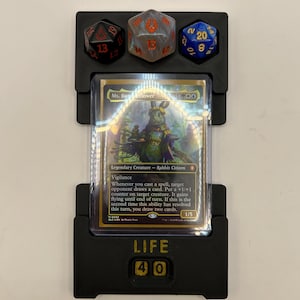 May include: Black 3D-printed card holder with dice slots and a life counter. The holder displays a Magic: The Gathering card featuring a rabbit character. Three multi-sided dice are in the top slots, and the life counter reads "40".