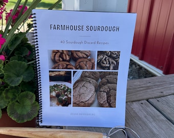 May include: A spiral-bound cookbook titled "Farmhouse Sourdough" with 40 sourdough discard recipes. A wooden handled whisk rests beside the book. The book features images of baked goods. The background includes a red building and greenery.