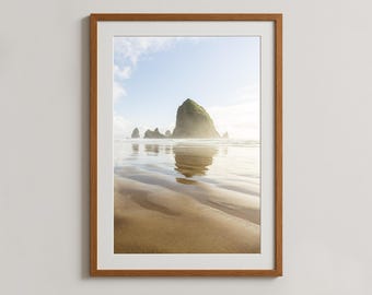 May include: Framed landscape photograph featuring a beach scene with a large rock formation in the ocean. The image captures the reflection of the rocks and sky on the wet sand. The sky is a light blue with some clouds, and the frame is a warm brown.