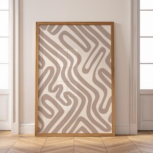Op de afbeelding: Een ingelijste abstracte kunstprint met een beige achtergrond en vloeiende, taupekleurige lijnen. Het kunstwerk is ingekaderd in een lichtbruine houten lijst, wat een minimalistische en moderne esthetiek creëert. De print wordt tegen een witte muur getoond.