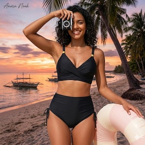 May include: A black two-piece swimsuit with a high-waisted bottom and a wrap-style top. The swimsuit has gold accents on the straps. The background shows a beach scene with boats and a sunset.