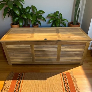 May include: A large, rectangular wooden storage chest with a light brown finish. The chest has a slatted front design and a flat top with a small, circular handle. The chest is on a wooden floor, with a decorative rug in front.