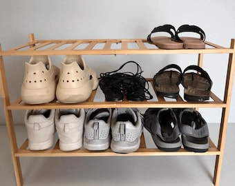May include: A three-tiered wooden shoe rack displaying various footwear. The rack holds cream-colored clogs, black sandals, and white sneakers. The rack is made of light-colored wood and is designed for home organization.