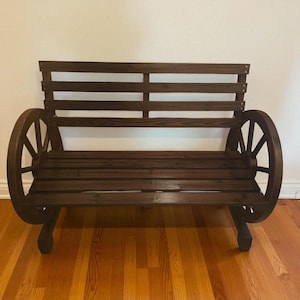 May include: A rustic wooden bench with a dark brown finish. The bench features a slatted seat and backrest, and is supported by large wagon wheel-style side supports. The bench is designed for outdoor or indoor use.