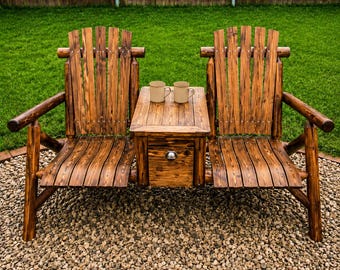 May include: A rustic wooden bench with two attached chairs and a small table in the middle. The bench has a drawer and two mugs on the table. The bench is made of natural wood and is set on a bed of small stones.