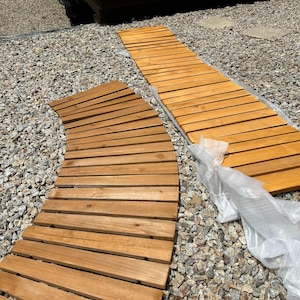 May include: Two wooden pathway mats, one curved and one straight, made of horizontal wooden slats. The mats are a warm, natural wood tone. They are resting on a bed of small, gray gravel. A piece of clear plastic is partially covering the straight mat.