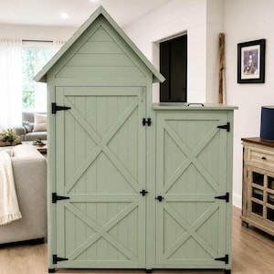 May include: A sage green wooden storage cabinet with a pitched roof design. The cabinet features two doors with an X-shaped cross design and black hinges. The cabinet has a small storage area on the side.