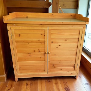 May include: A light brown wooden cabinet with two doors and a top shelf. The cabinet is made of horizontal wooden planks and has two round wooden knobs. The cabinet is sitting on a wooden floor.